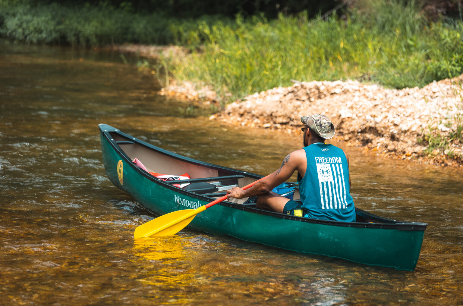 A man solo canoeing down the Huzzah River surrounded by lush green Ozark forest.